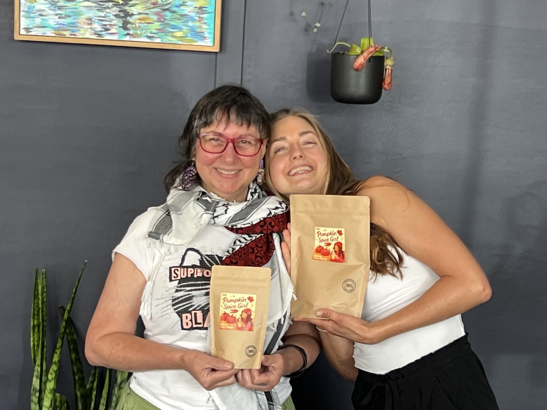 Emilie Meyer and Nada Todorovich, co-founders of Pumpkin Spice Girl, each holding up the packaged Pumpkin Spice Girl blend, smiling at the camera. Emilie has her head on Nada's shoulder.